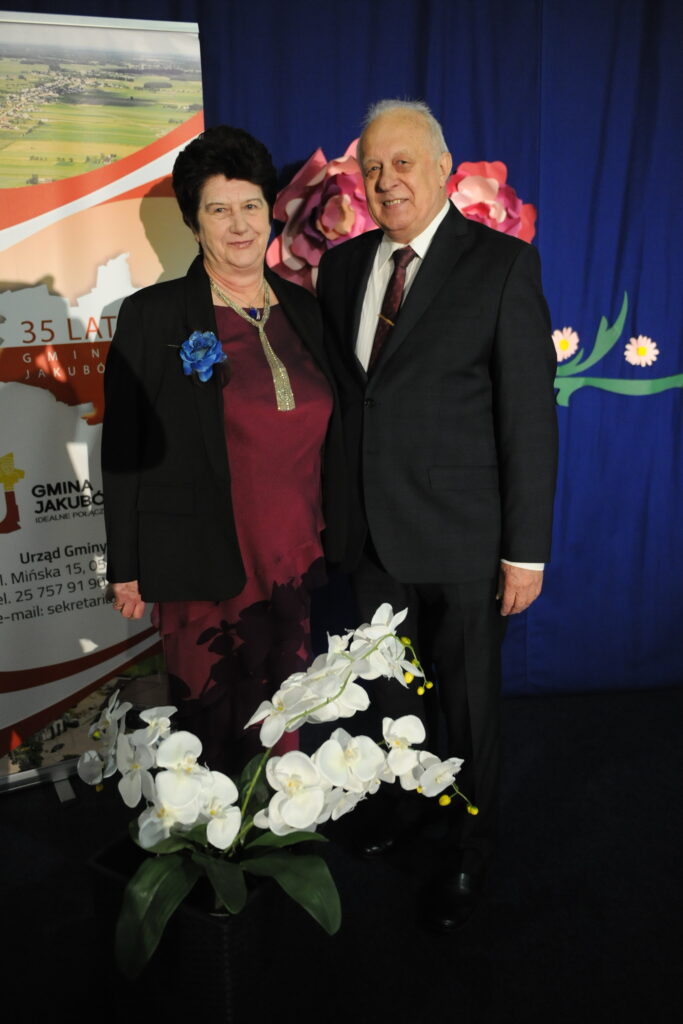 Starsze osoby stoją na tle niebieskiej zasłony z kwiatami. Pani ma bordowy strój z niebieską broszką, pan w garniturze. Z przodu białe orchidee w doniczce.