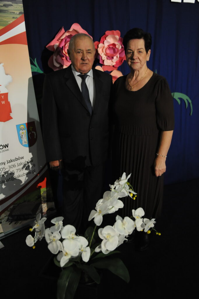 Na zdjęciu para w eleganckich strojach stoi przed ciemnoniebieskim tłem. Widoczne są dekoracyjne, różowe kwiaty i biały plakat. Na pierwszym planie białe orchidee dodają kontrastu.