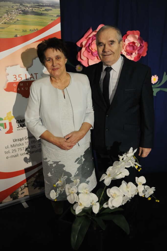 Para elegancko ubranych osób stoi przed niebieskim tłem z dekoracjami kwiatowymi. Obok widać biały plakat z czerwonymi akcentami i orkideę. atmosfera jest formalna i uroczysta.