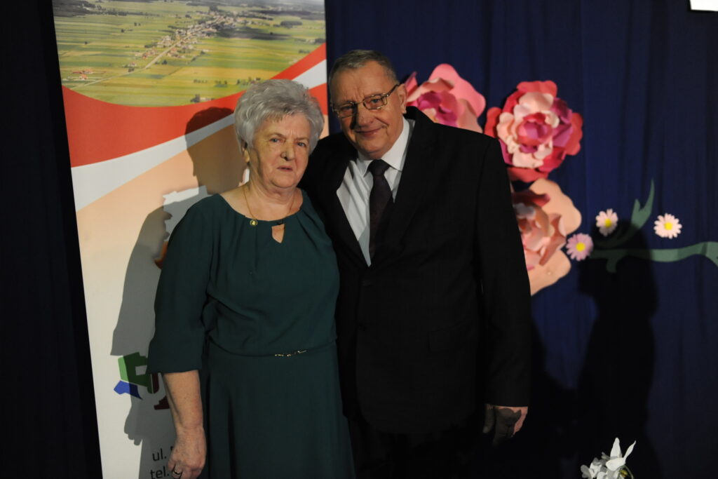 Na zdjęciu starsza kobieta i mężczyzna w eleganckich strojach, na tle niebieskiej dekoracji z kwiatami. Kobieta ma zieloną sukienkę, a mężczyzna ciemny garnitur. Obok wiszą banery z krajobrazem.
