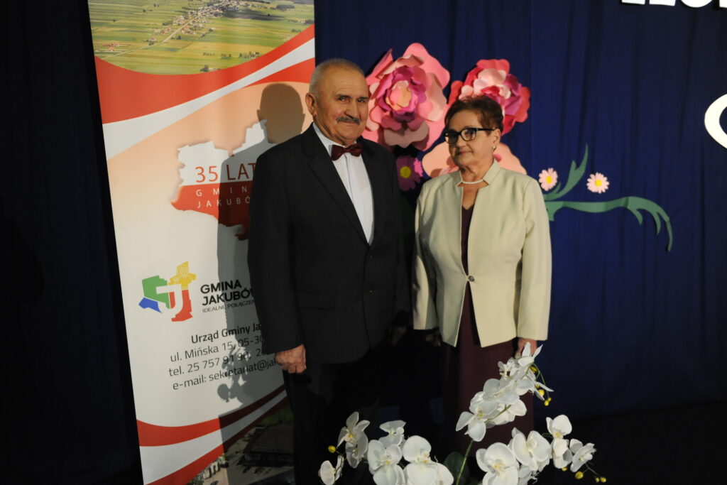 Dwoje ludzi stoi obok plakatu z napisem Gmina Jakubów. Tło jest granatowe, ozdobione kwiatami. Na pierwszym planie białe orchidee.