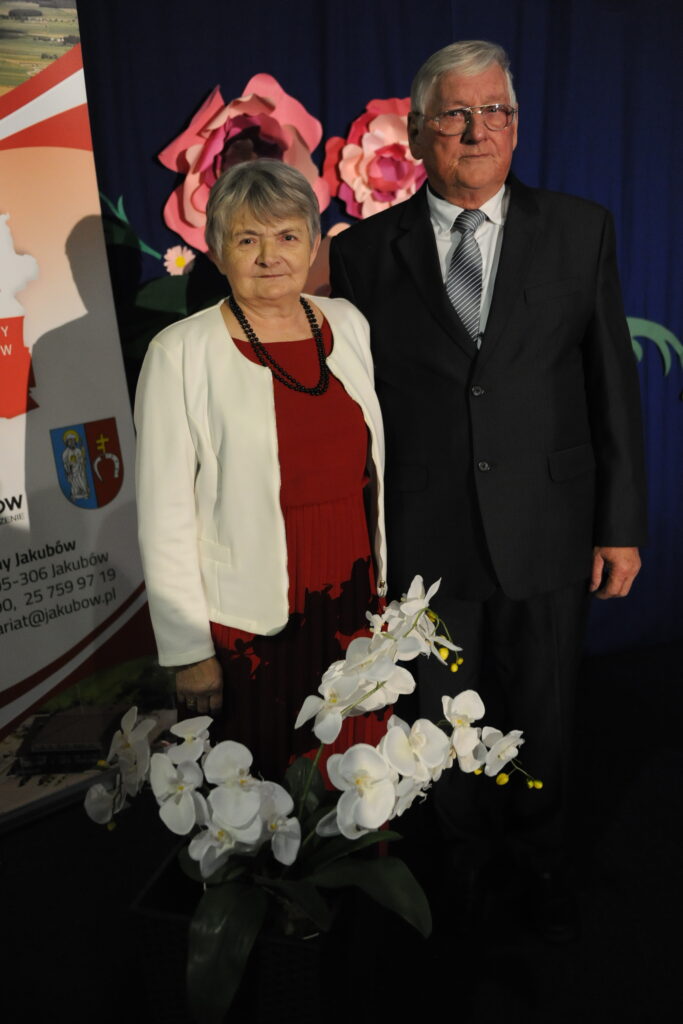 Para starszych osób stoi przed ciemnym tłem z kwiatami. Kobieta ma białą marynarkę, a mężczyzna ciemny garnitur. Na pierwszym planie białe orchidee.