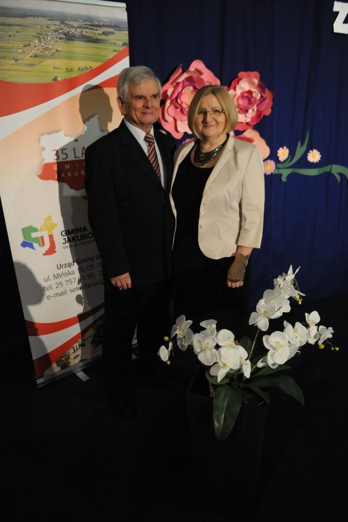 Para w eleganckich strojach stoi na tle niebieskiej kurtyny z kwiatami. Po lewej baner z napisem 35 lat. Przed nimi białe orchidee.