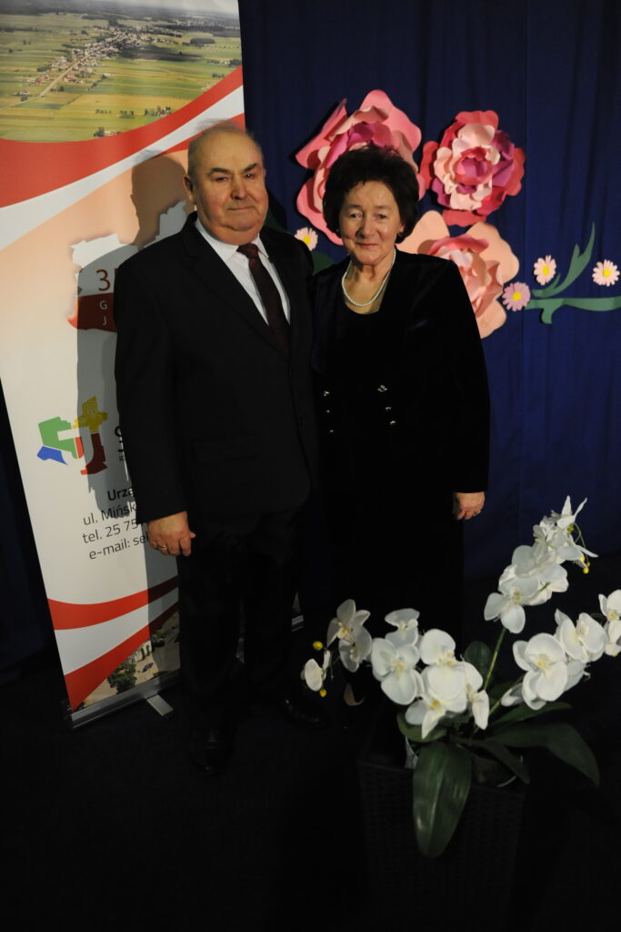 Para stoi przed niebieskim tłem z różowymi kwiatami. Mężczyzna w garniturze i kobieta w czarnej sukni. Białe orchidee na pierwszym planie.