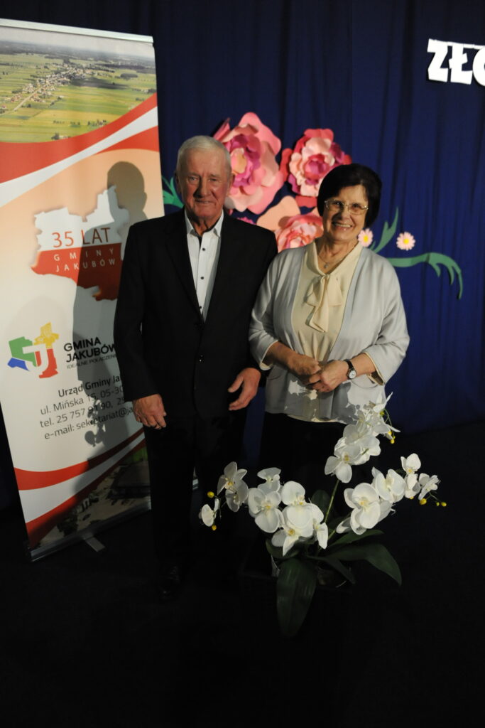 Para starszych osób stoi przed banerem z napisem 35 lat gminy Jakubów. Tło zdobią różowe kwiaty na granatowym tle. Na pierwszym planie znajduje się biała orchidea.