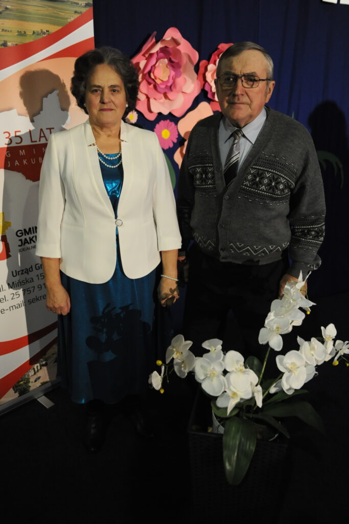 Dwie osoby stoją na tle niebieskiej kurtyny z kwiatowym dekorem, kobieta w białej marynarce, mężczyzna w szarym swetrze. Po ich lewej baner z informacjami. Na pierwszym planie białe orchidee.