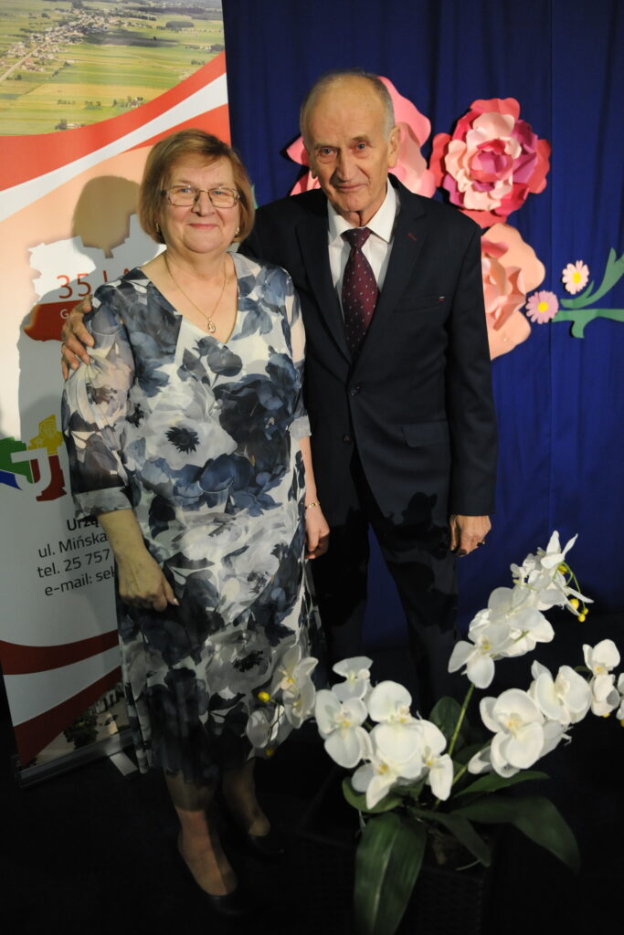 Starsza para pozuje na tle niebieskiej zasłony z papierowymi kwiatami. Kobieta ma kwiecistą sukienkę, mężczyzna ciemny garnitur. Na pierwszym planie białe storczyki.