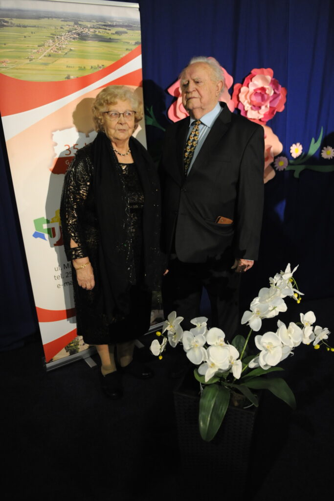 Para starszych ludzi stoi przy plakacie na tle z kwiatami. Kobieta w czerni, mężczyzna w garniturze. Przód zdobią białe orchidee.