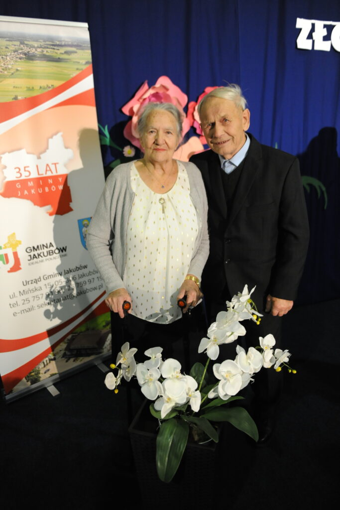 Starsza para stoi przed niebieską zasłoną, trzymając się za ręce. Obok nich biały plakat i donica z białymi orchideami. W tle widoczne są papierowe kwiaty.