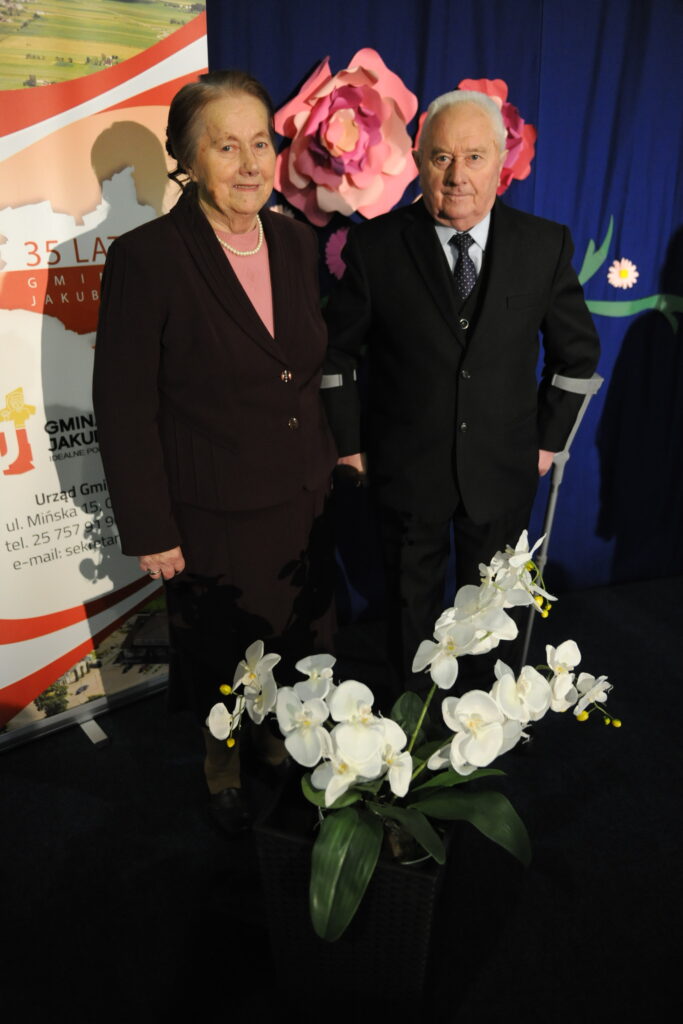 Dwójka starszych osób w eleganckich strojach stoi przed niebieskim tłem i kwiatami. Obok nich stoi plakat z napisem. Na pierwszym planie białe orchidee.