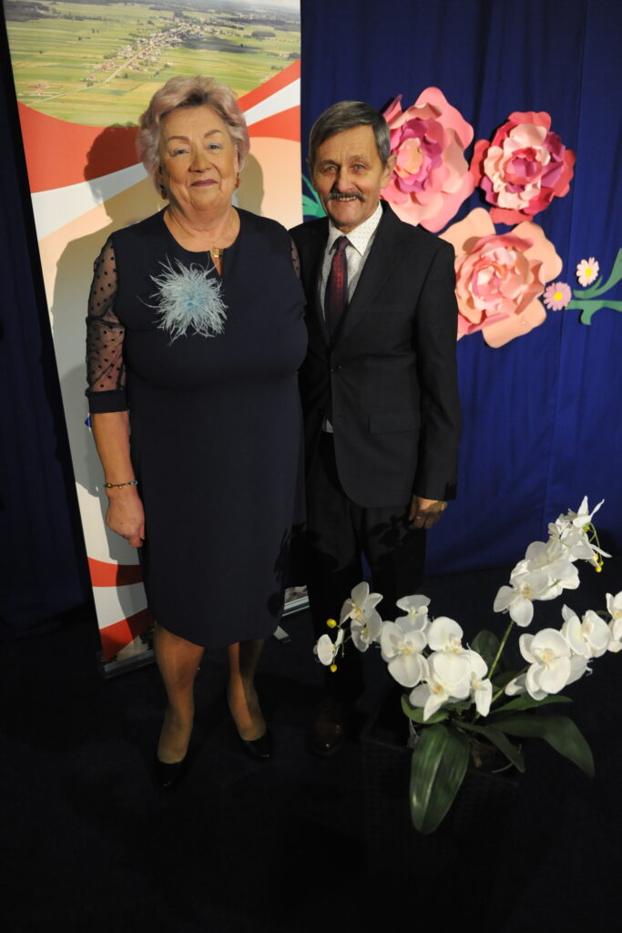 Na zdjęciu starsza kobieta i mężczyzna stoją razem na tle granatowej zasłony z różowymi kwiatami. Kobieta ma granatową sukienkę z błękitnym ozdobieniem, a mężczyzna ciemny garnitur z krawatem. Na pierwszym planie biały buk