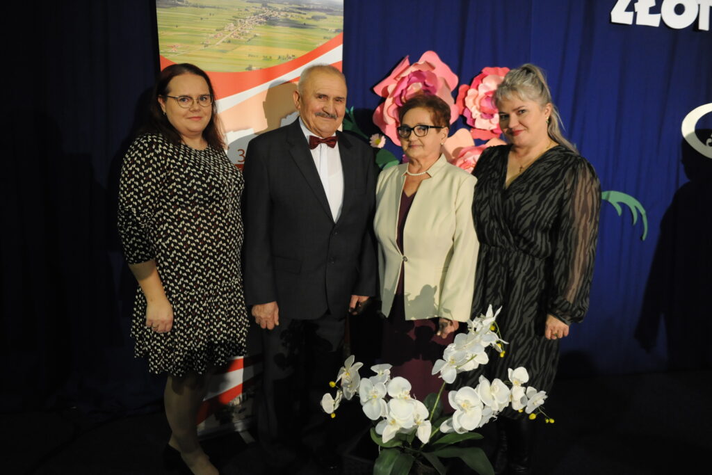 Cztery osoby stoją na tle niebieskiej ściany z różowymi kwiatami i plakatem. Mają na sobie eleganckie stroje. Na pierwszym planie widać białe orchidee.