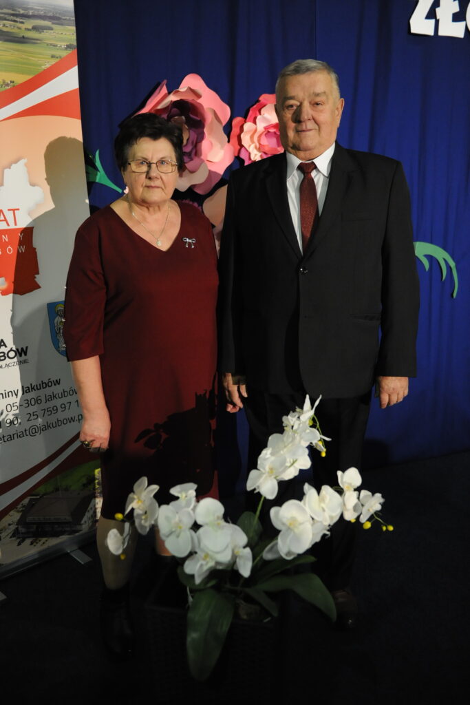 Na zdjęciu widać dwie osoby w eleganckich ubraniach, tle ozdobione kwiatami, a obok biała orchidea.