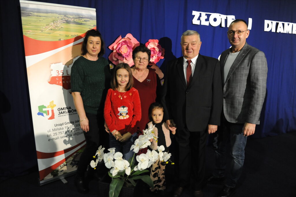 Sześć osób stoi przed niebieskim tłem z napisem. Przy nich są kwiaty i roll-up z logiem Gminy Jakubów.