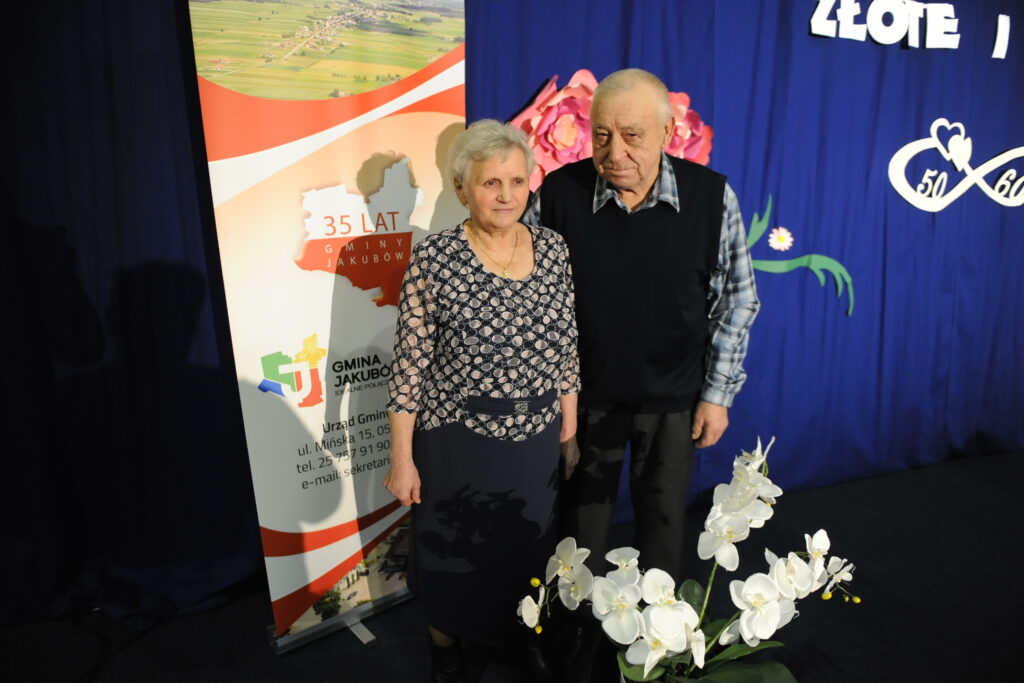 Starsza para pozuje na tle niebieskiego materiału. Obok nich baner gminy Jakubów. Przed nimi białe orchidee.