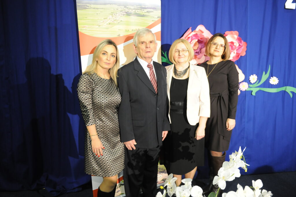 Cztery osoby pozują na tle niebieskiej zasłony. Z tyłu widać kwiaty i plakat z krajobrazem. Na pierwszym planie białe kwiaty.