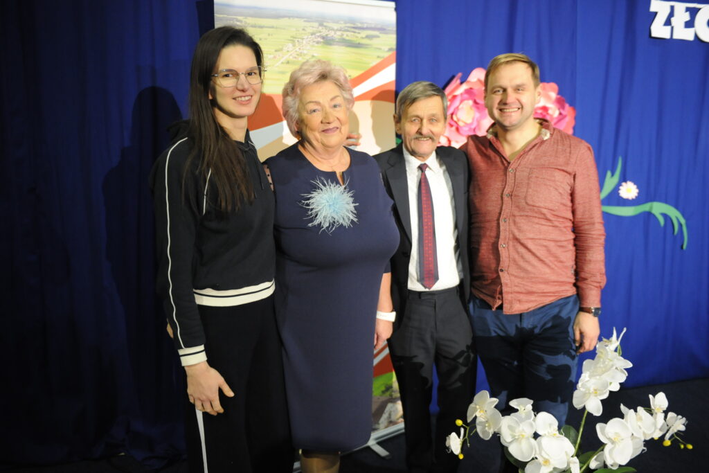 Na zdjęciu są cztery uśmiechnięte osoby na niebieskim tle. Kobieta w środku nosi ciemnoniebieską sukienkę z niebieską broszką. Przed nimi stoi wazon z białymi kwiatami.