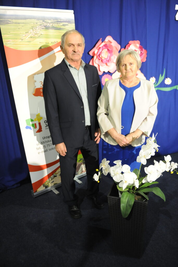 Dwie osoby w formalnym stroju stoją obok siebie. Tło jest niebieskie z dekoracyjnymi kwiatami. Na pierwszym planie biały storczyk.