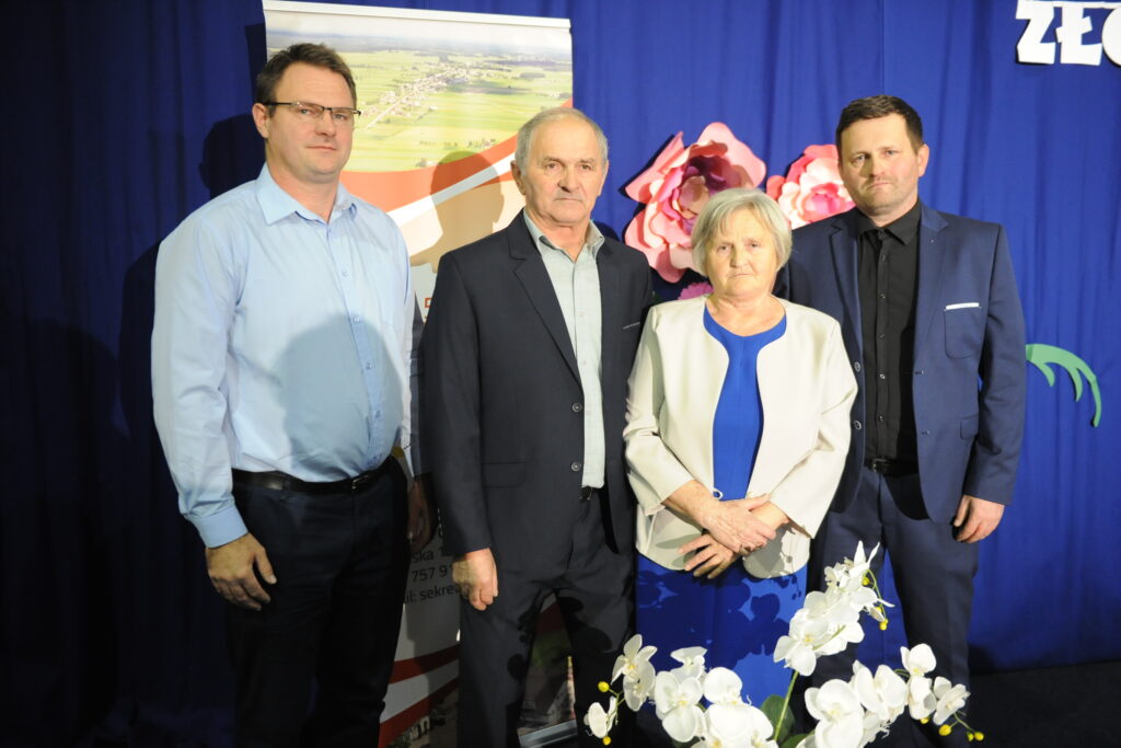 Na zdjęciu są cztery osoby w eleganckich ubraniach, stojące na tle niebieskiej zasłony. W tle widoczny jest plakat z krajobrazem i papierowe kwiaty. Na pierwszym planie znajdują się białe orchidee.