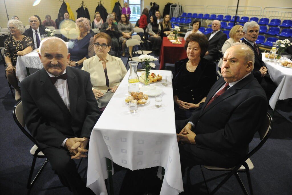 Na zdjęciu jest grupa osób siedzących przy stole z białym obrusem. Jedzą przekąski w jasnym pomieszczeniu z niebieskimi krzesłami w tle. Ubrani są elegancko, co sugeruje formalną okazję.
