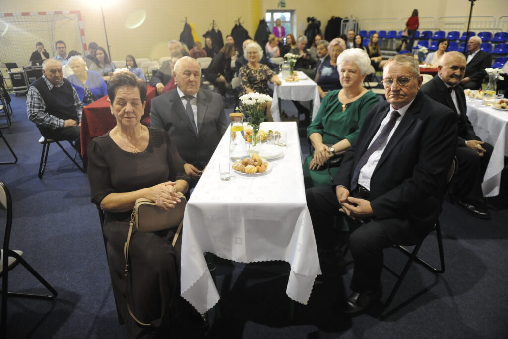 Ludzie siedzą przy stole na uroczystości w świetlicy. Stół przykryty białym obrusem, na nim ciastka i kwiaty. W tle niebieskie krzesła i żółte ściany.