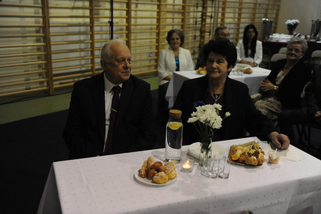 Starsza para siedzi przy białym stole na przyjęciu, obok talerzy z jedzeniem. Na stole ozdobione kwiaty i świeca. W tle inne osoby i naczynia.