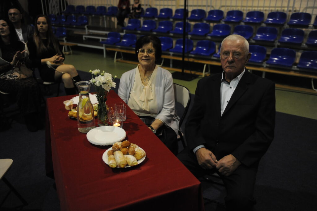 Para starszych ludzi siedzi przy stole nakrytym czerwonym obrusem z kwiatami i jedzeniem. W tle niebieskie krzesła na widowni. Oboje elegancko ubrani.