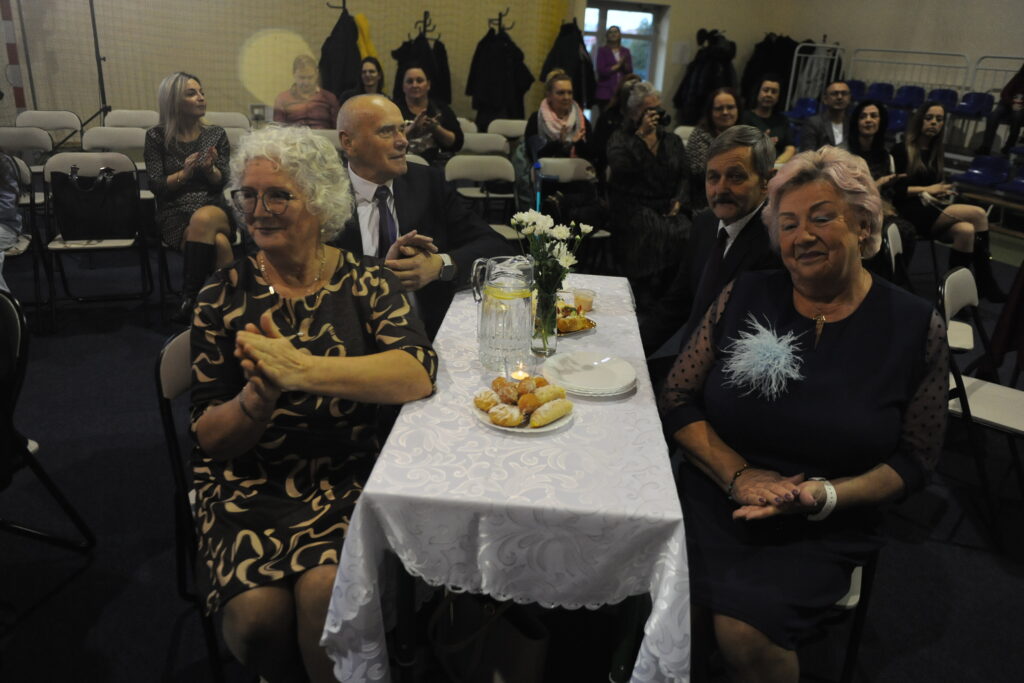 Grupa osób siedzi przy stole nakrytym białym obrusem, na którym stoją dzbanek i talerze z ciastkami. W tle znajduje się więcej osób siedzących w sali z wieszakami na płaszcze. Dominuje ciemne tło z jasnymi akcentami ubr