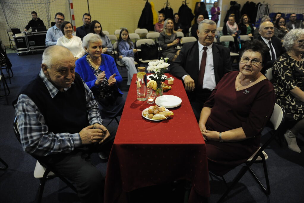 Starsze osoby siedzą przy stole z czerwonym obrusem, na którym leżą przekąski. W tle grupa ludzi na widowni. Ciemne wnętrze pomieszczenia.