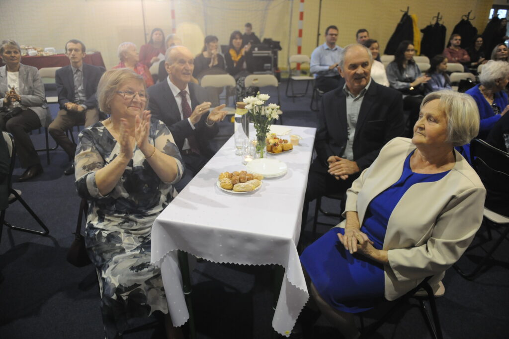 Grupa osób przy stole z białym obrusem i poczęstunkiem. W tle widać ludzi siedzących na krzesłach. Atmosfera wygląda na uroczystą, wszyscy są elegancko ubrani.