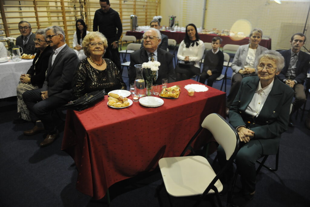 Grupa osób siedzi przy stole z czerwonym obrusem, na którym są przekąski. Pomieszczenie z jasnym oświetleniem i drewnianymi listwami na ścianie. W tle ludzie siedzą przy stole z białym obrusem.