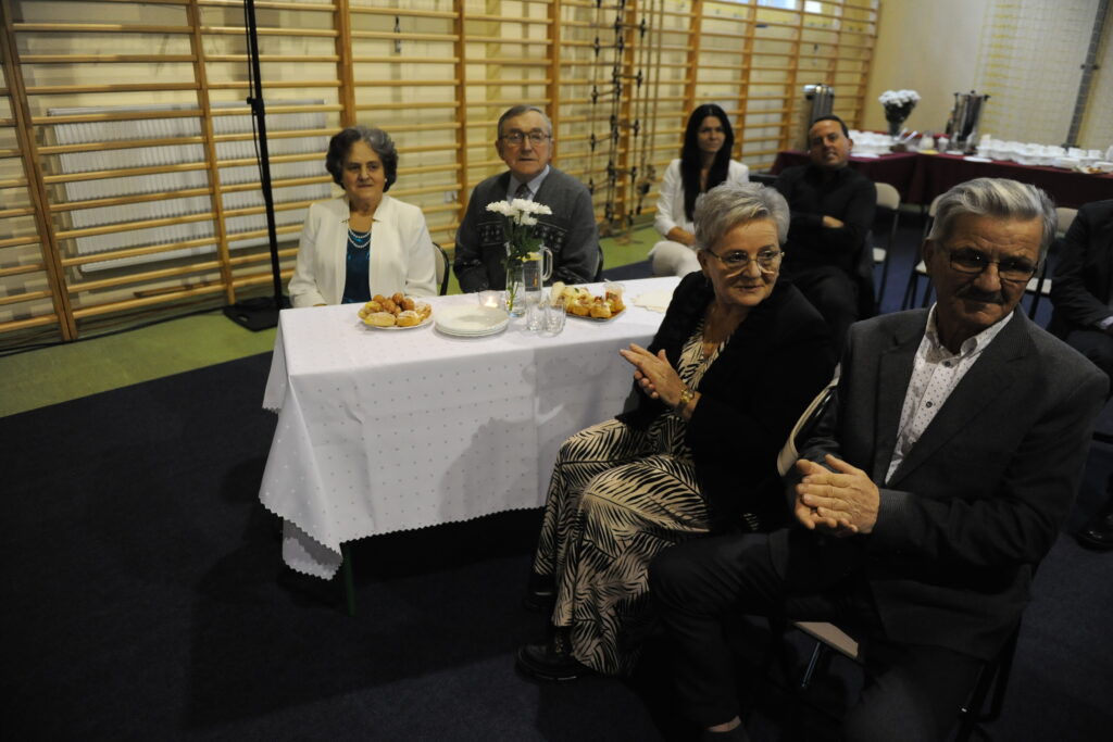 Grupa starszych osób siedzi przy stole z białym obrusem, na którym są ciastka i kwiaty. Tło to jasna sala z drewnianymi drabinkami. Ubioru dominują eleganckie stroje w neutralnych kolorach.