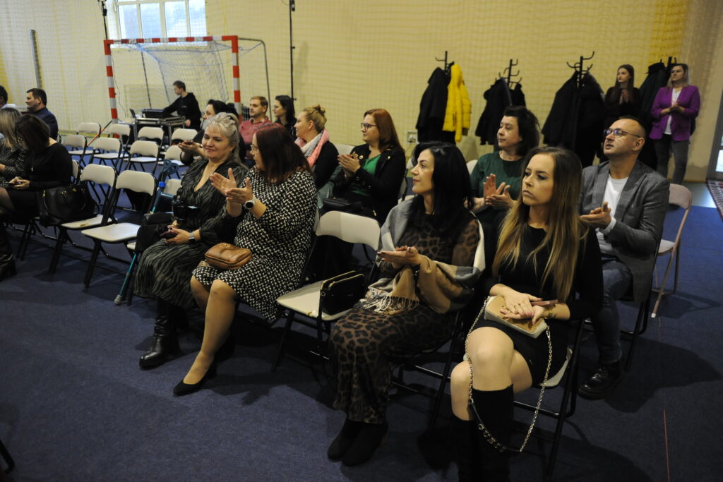 Grupa ludzi siedzi na krzesłach w sali z niebieską wykładziną. W tle widać wieszaki i bramkę do gry. Uczestnicy biją brawo, ubrani w ciemne kolory.