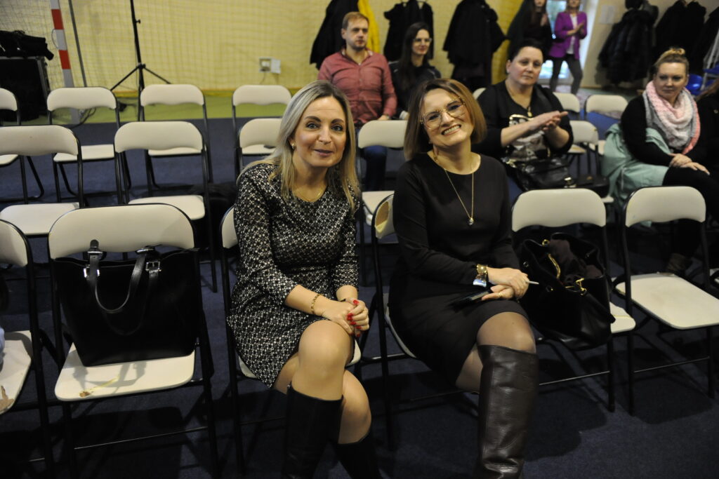 Dwie kobiety siedzą na krzesłach w sali; jedna w sukience w czarne wzory, druga w czarnej sukience. W tle puste krzesła i wiszące płaszcze. Podłoga jest pokryta ciemnym dywanem.