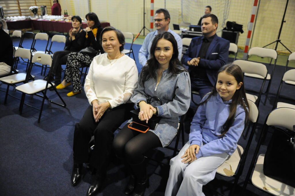 Na obrazie widoczne są trzy osoby siedzące na krzesłach, w tym jedna dziewczynka. Tło jest dobrze oświetlone z widoczną siatką sportową. Przeważają kolory ciemnoniebieski i jasne odcienie ubrań.