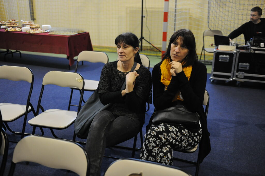 Dwie kobiety siedzą na krzesłach w sali z niebieską wykładziną. W tle stół z czerwonym obrusem i jedzeniem. Obok sprzęt audio, a za nimi siatka sportowa.