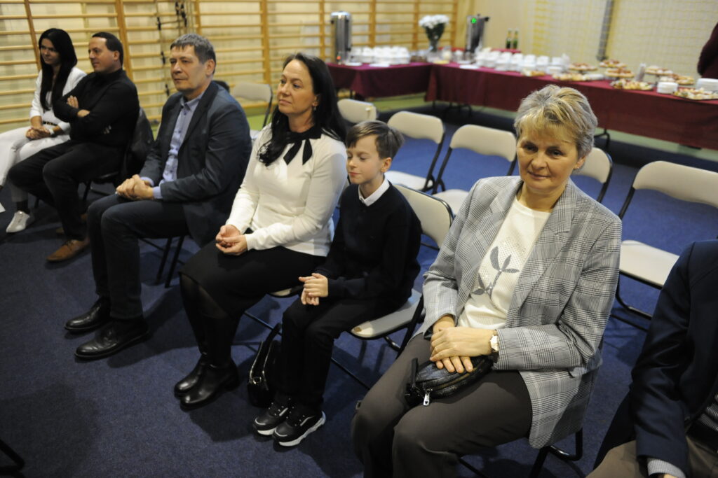 Grupa ludzi siedzi na krzesłach, tło z jasnymi ścianami i stołem z przekąskami. Kobieta w szarej marynarce, chłopiec w czerni. Atmosfera formalna.