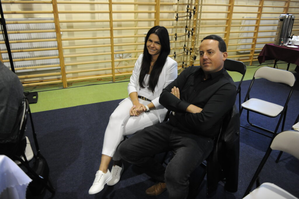 Mężczyzna i kobieta siedzą na krzesłach, ubrani w czarne i białe stroje. W tle jasne drewniane drabinki gimnastyczne. Na podłodze zielono-niebieski dywan.