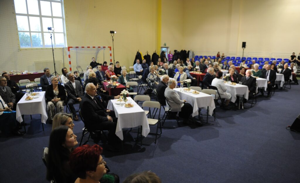 Sala wypełniona ludźmi siedzącymi przy stołach z białymi obrusami. Ściany jasnożółte, wyłożone krzesłami i sprzętem sportowym. W centrum uwagi osoby w eleganckich strojach, atmosfera uroczysta.