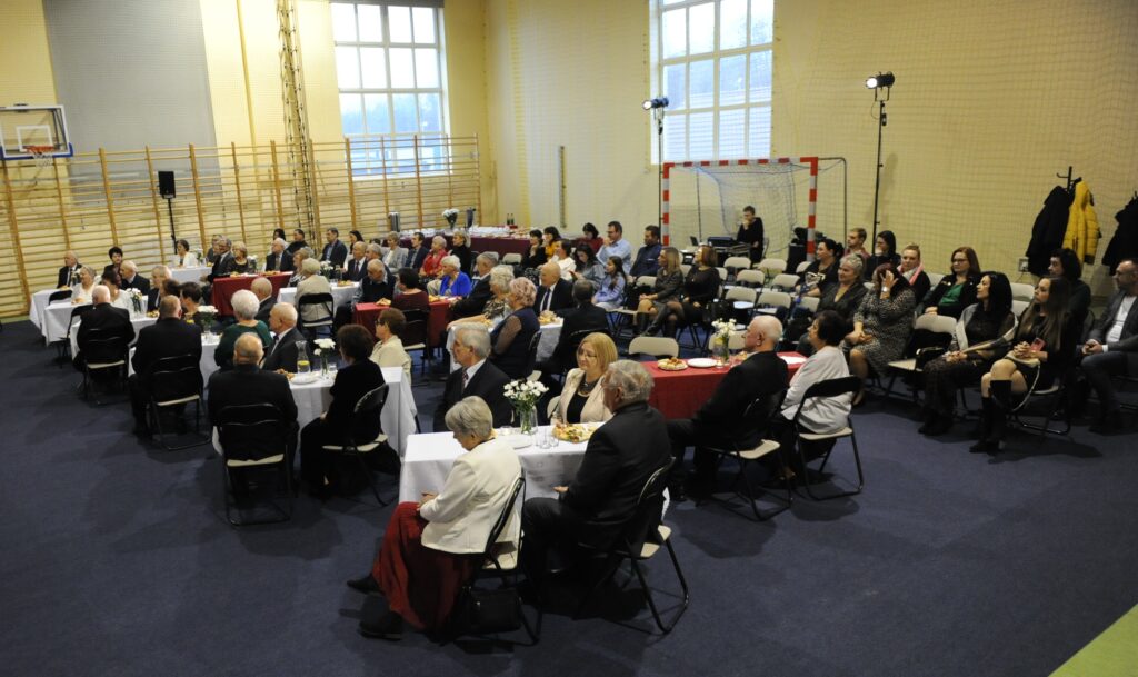 Na zdjęciu widać ludzi przy stołach w sali gimnastycznej. Dominują barwy białe i czerwone, z niebieską podłogą. W tle są duże okna i drabinki.