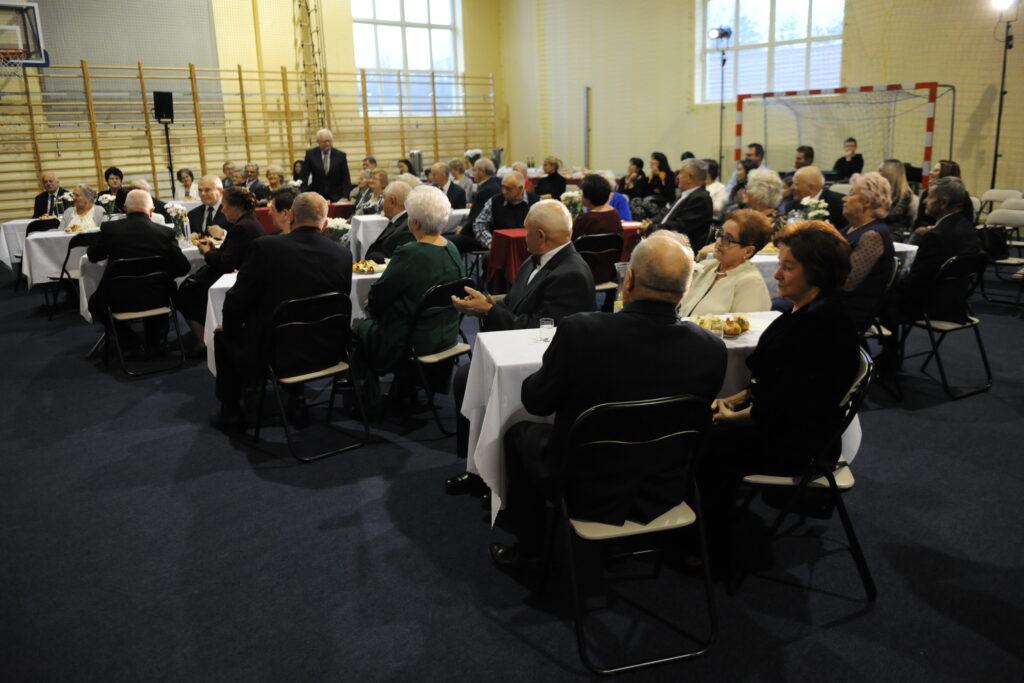 Grupa starszych osób siedzi przy stołach w hali sportowej. Ubrani w eleganckie stroje, na stołach białe obrusy. Tło stanowią drabinki gimnastyczne i bramka.