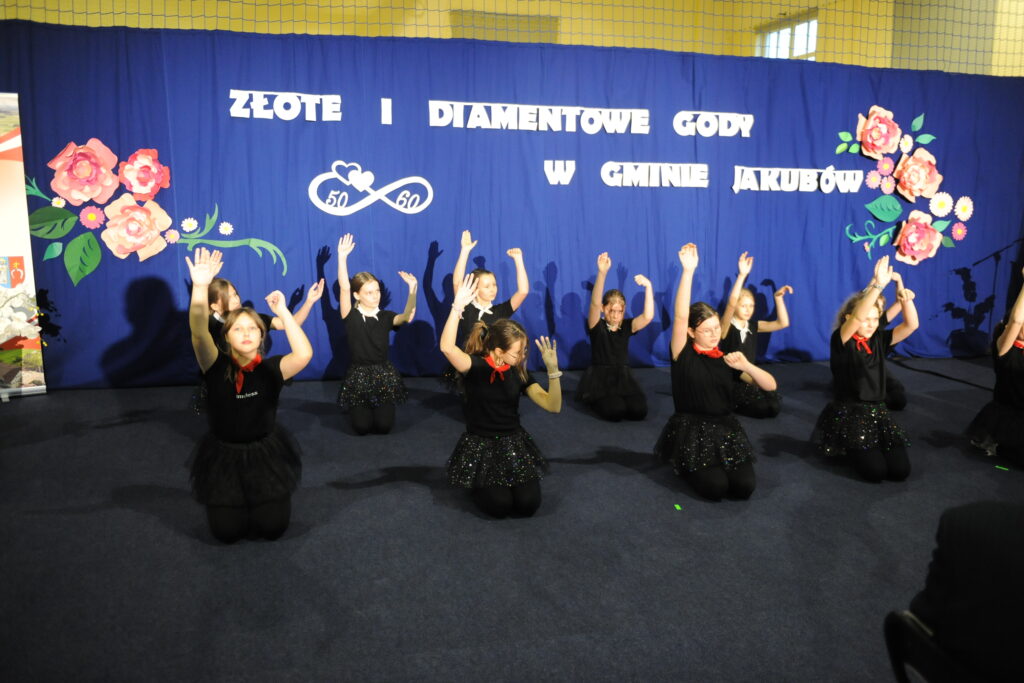 Grupa młodych osób w czarnych strojach tańczy na scenie. Tło to niebieska kurtyna z napisami i dekoracjami kwiatowymi. Wydarzenie nosi tytuł Złote i Diamentowe Gody.