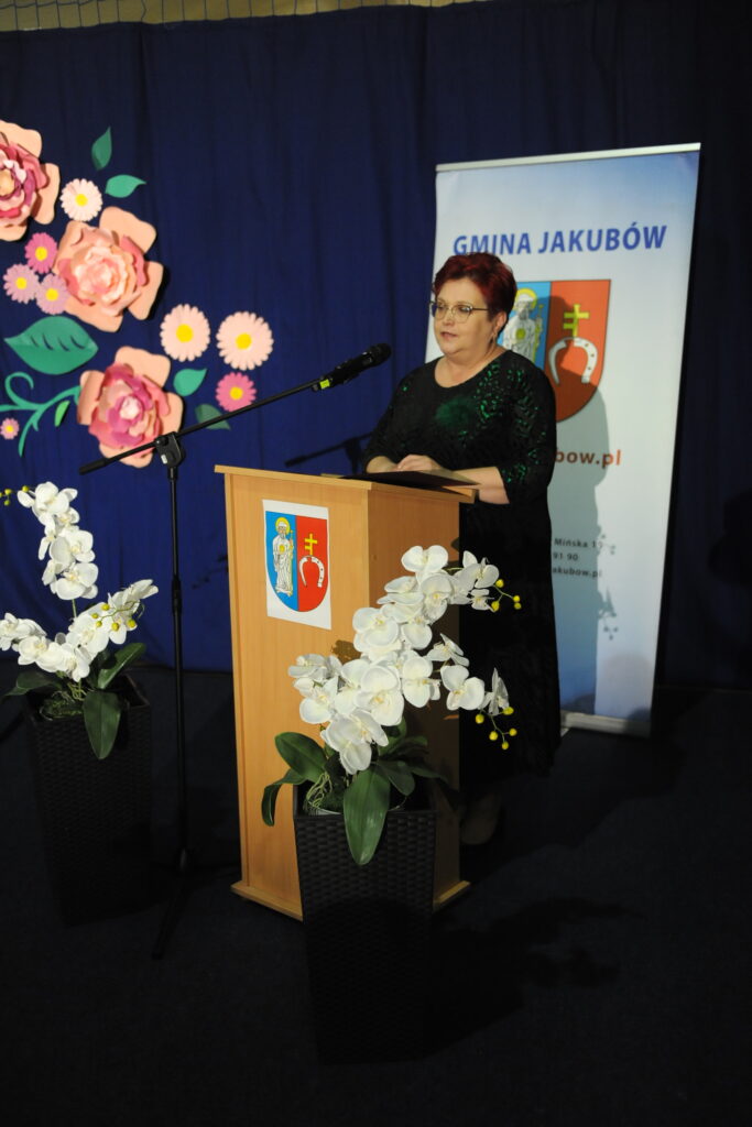 Osoba przy mównicy na tle niebieskiej zasłony z papierowymi kwiatami. Obok stoją dwie donice z białymi orchideami. Po prawej baner Gminy Jakubów.