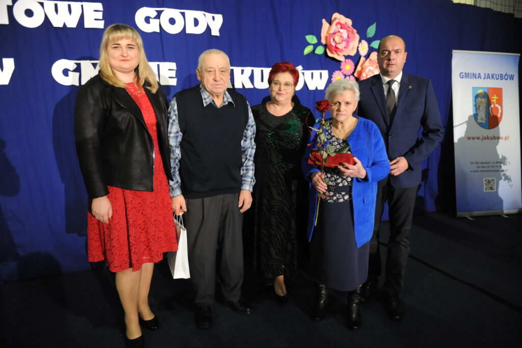 Pięć osób stoi na tle niebieskiej ściany z kwiatami i napisami. Jedna osoba trzyma różę. Obok nich znajduje się banner gminy Jakubów.