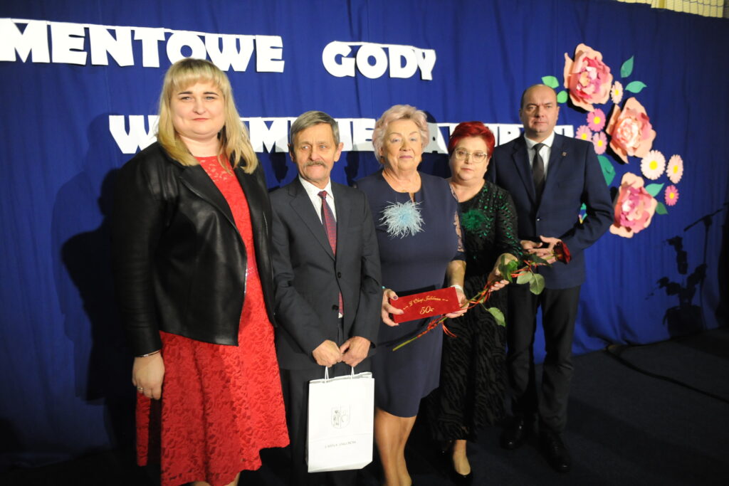 Pięć osób stoi przed niebieskim tłem z napisami i kwiatami. Dwie osoby trzymają róże i tabliczkę. Grupa ubrana elegancko.