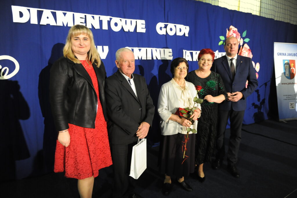 Pięć osób stoi na tle ciemnoniebieskiego banera z napisem. Dwie kobiety trzymają róże. Ciemne ubrania dominują.