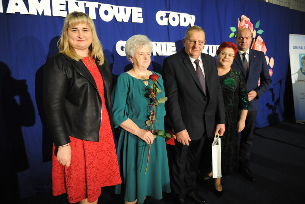 Pięć osób na tle niebieskiego banera z napisami. Kobieta w zielonej sukni trzyma różę. Eleganckie stroje, uroczysta atmosfera.