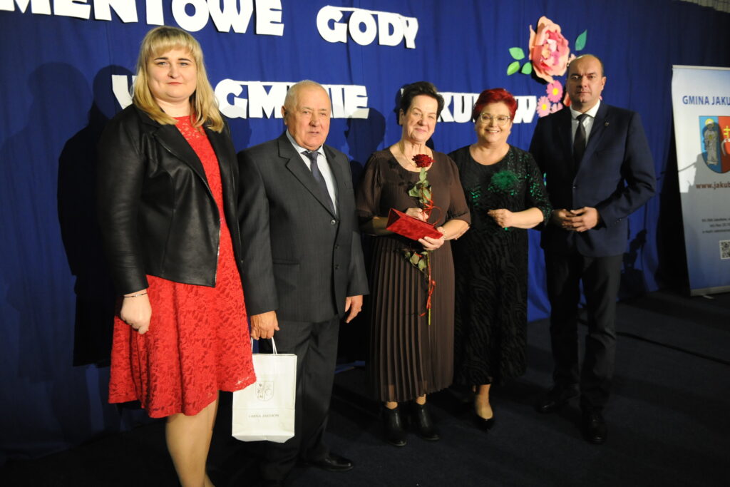 Na zdjęciu pięć osób stoi przed niebieskim tłem z białym napisem. Kobieta w czerwonej sukience trzyma białą torbę. Starsza kobieta z różą w dłoni pośrodku grupy.