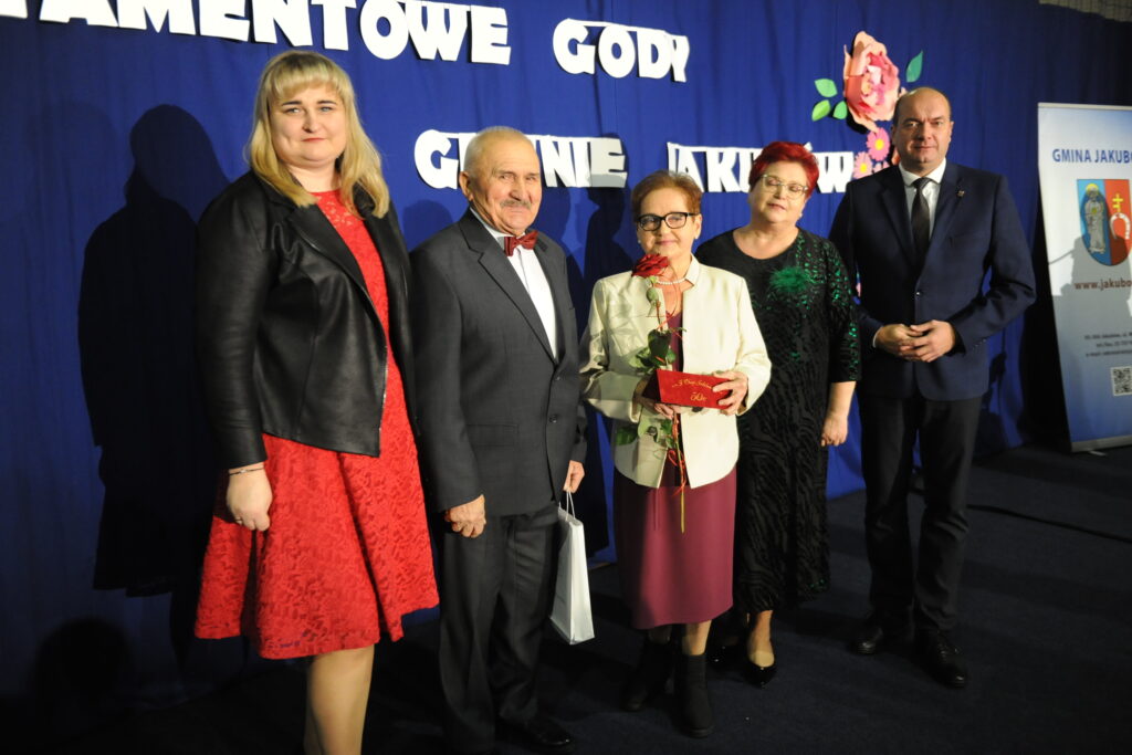 Pięć osób stoi przed niebieskim tłem z napisem. Kobieta w czerwonej sukience trzyma prezent. Obok niej mężczyzna w garniturze.