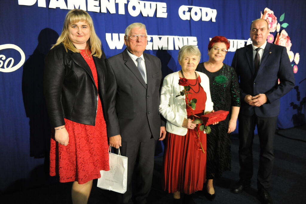 Pięć osób stoi razem na tle granatowego banera z napisem. Dwie kobiety w czerwonych sukienkach, mężczyźni w garniturach. Jedna osoba trzyma różę.